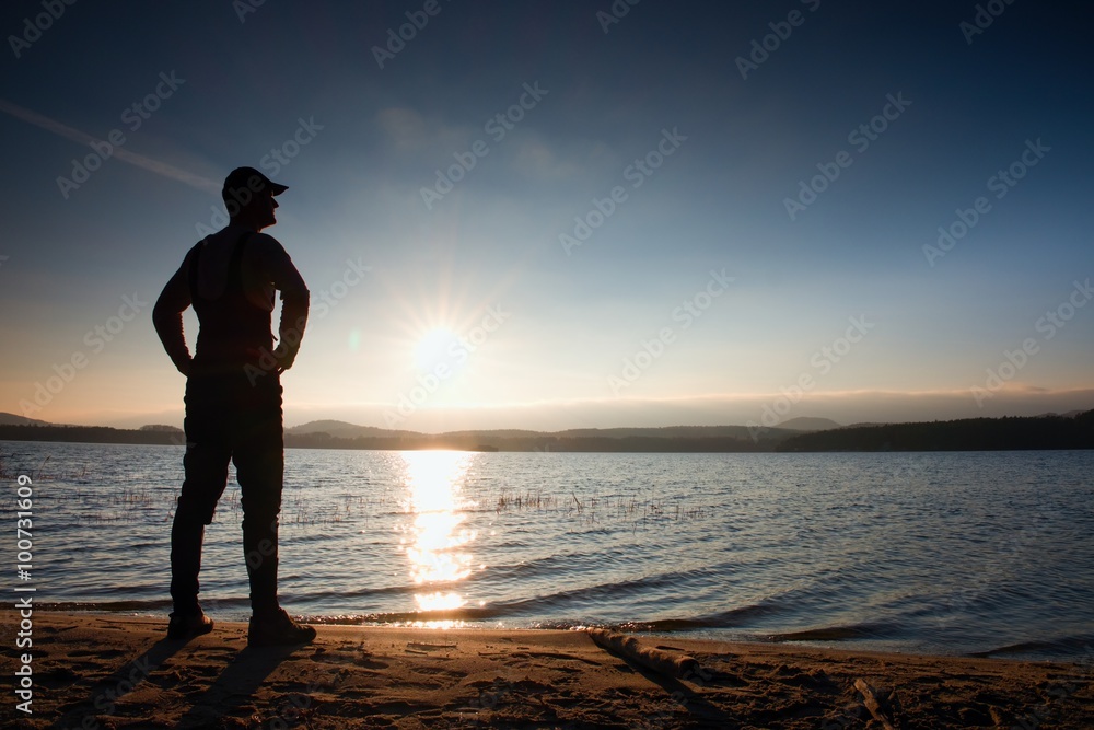Tall hiker in dark sportswear with poles and sporty backpack on beach enjoy sunset at horizon with blue sky with clouds.