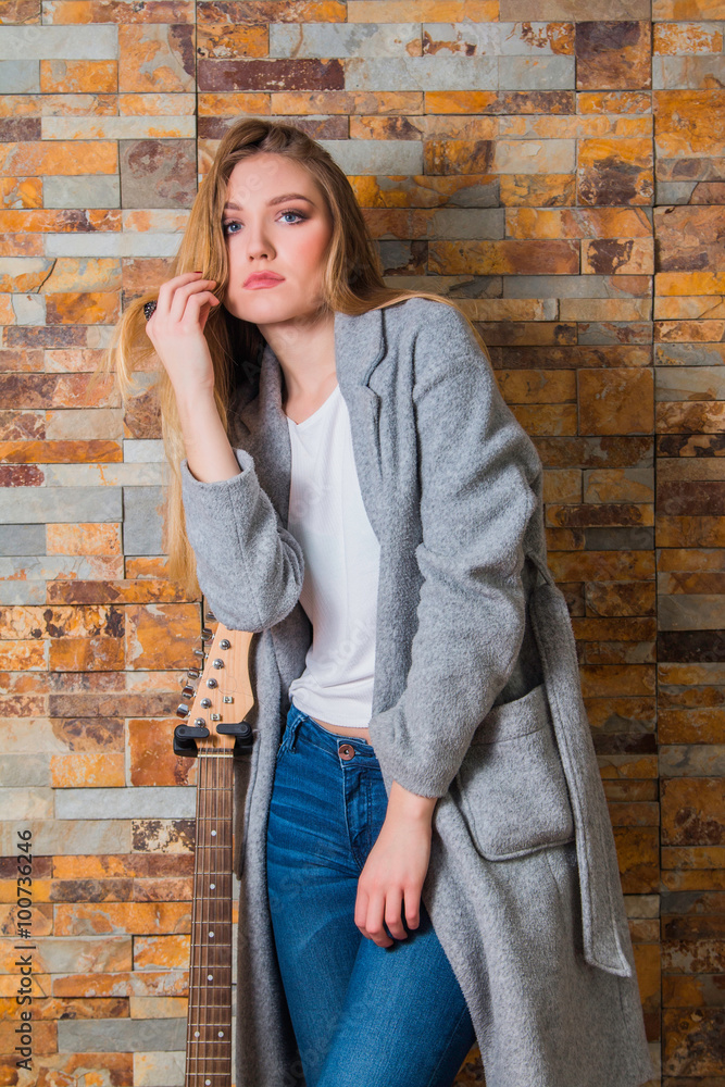 Beautiful young girl in overcoat and jeans posing with guitar on brick background Beautiful young girl in overcoat and jeans posing with guitar on brick background
