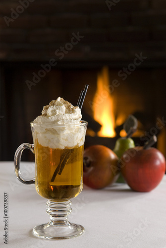 Hot apple cider drink topped with whipped cream and cinnamon. Fireplace and apples in the background.