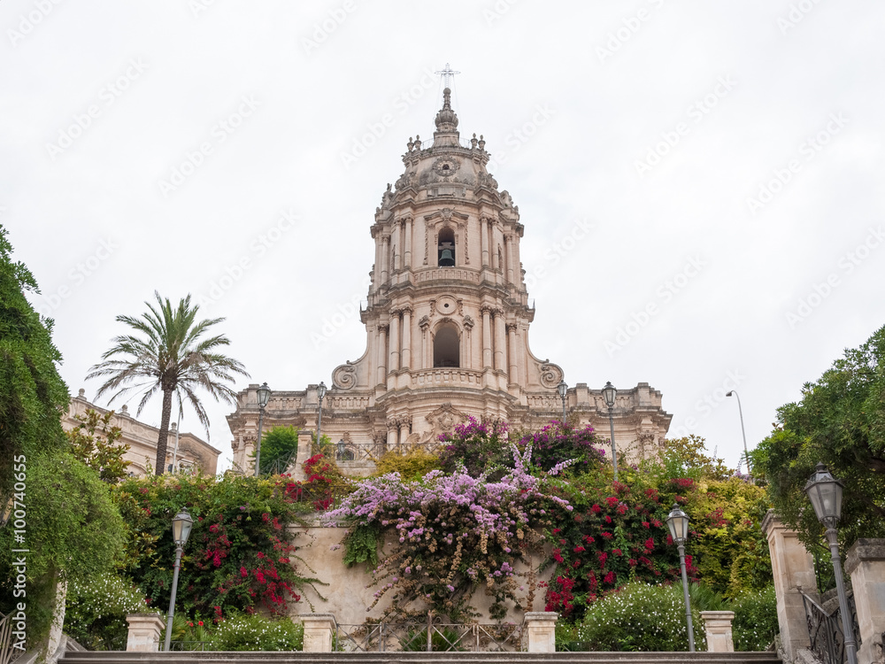 The baroque facade of the Modica's cathedral with several flowers in the foreground