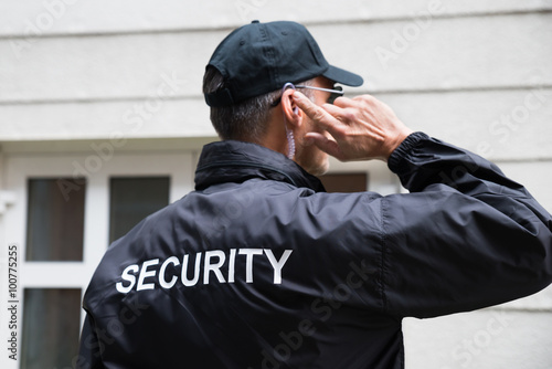 Fotografia, Obraz Security Guard Listening To Earpiece Against Building