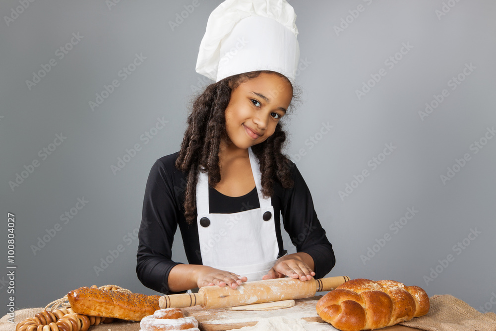 Little dark-skinned girl rolls the dough. The child learns to cook. Clothing and chef hat Little dark-skinned girl rolls the dough. The child learns to cook. Clothing and chef hat