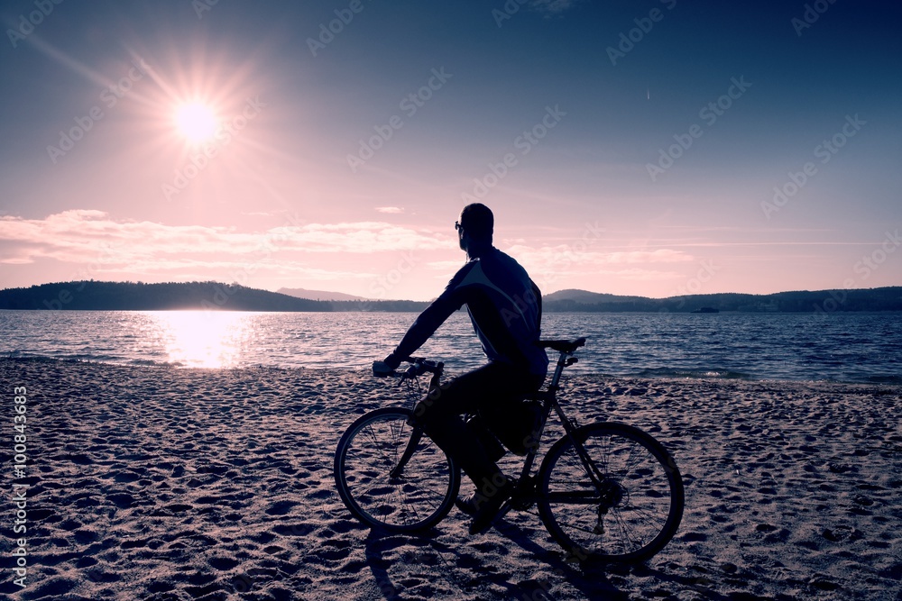 Fototapeta premium Young man cyclist silhouette on blue sky and sunset background on the beach. End of season at lake.