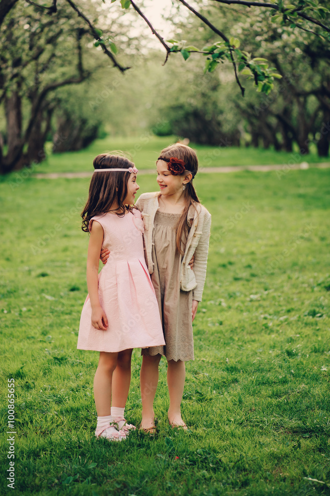 two happy dressy little girlfriends hugs outdoor in spring garden. Kid sisters spending time together on the walk - obrazy, fototapety, plakaty 