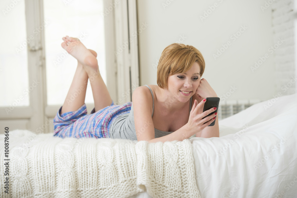 young attractive girl with red hair internet networking with mobile phone lying on bed in sleeping pants young attractive girl with red hair internet networking with mobile phone lying on bed in sleeping pants