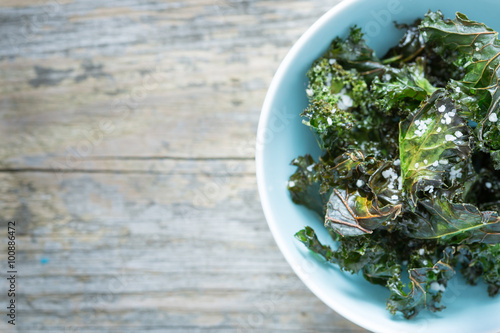 Kale chips with salt in pastel blue bowl captured from above  top view . Healthy dietetic snack. Vintage rustic wood as background. Background layout with free text space.