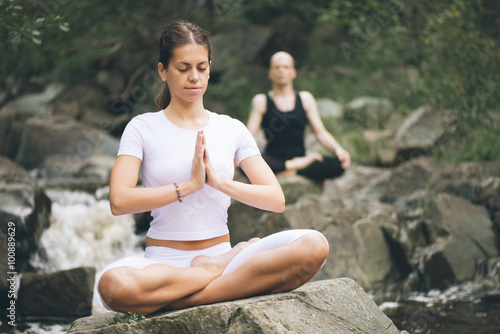 Beautiful young woman with a man behind meditating in yoga pose at a mountain stream. Selective focus on woman.