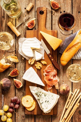 Fotografía Different kinds of cheeses, wine, baguette, fruits and snacks on rustic wooden table from above