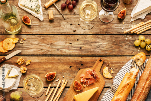 Different kinds of cheeses, wine, baguettes, fruits and snacks on rustic wooden table from above. French tasting party or feast scenery. Background layout with free text space.