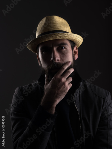 Thinking bearded man wearing straw hat touching beard looking at camera. High contrast low key dark shadow portrait over black background.