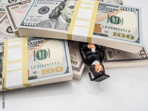 Photograph of figurine of unhappy female graduate in cap and gown buried under stacks of U.S. paper currency, suggesting the idea of the high price of education and crushing student loans.