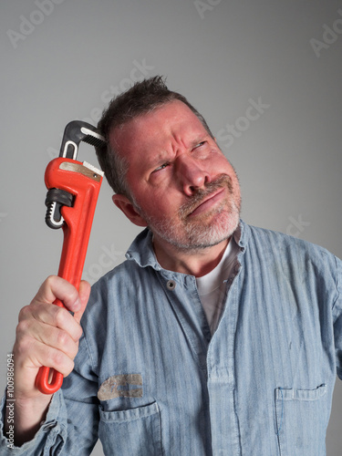 Photograph of a repairman in blue coveralls looking confused and scratching his head with an orange pipe wrench (humor).