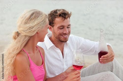 Happy young couple having red wine outdoors on beach. Smiling man and woman in love are holding bottle and wineglasses. Loving male and female are in casual clothing.
