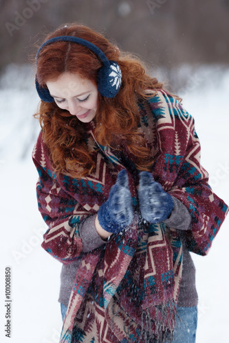 Portrait of attractive young curly red-haired woman in blue gloves and headphones. She is wrappening in a big scarf with ethnic pattern. She claps her hands and enjoys the snow.