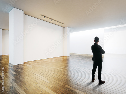 Business man stands opposite white wall in museum interior with wooden floor. Horizontal