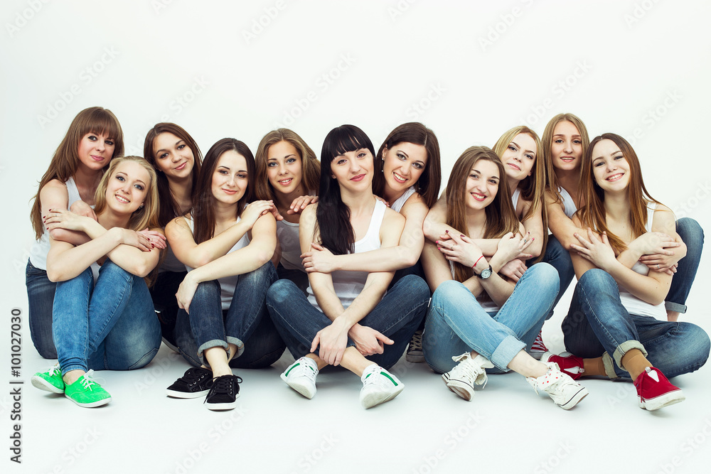 Obraz premium Happy together concept. Group portrait of healthy girls in white t-shirts and blue jeans sitting and posing over white background. Copy-space. Urban style. Studio shot