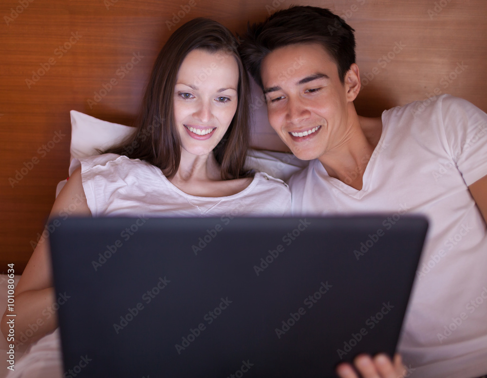young couple sitting on a bed with happy smile, using laptop.