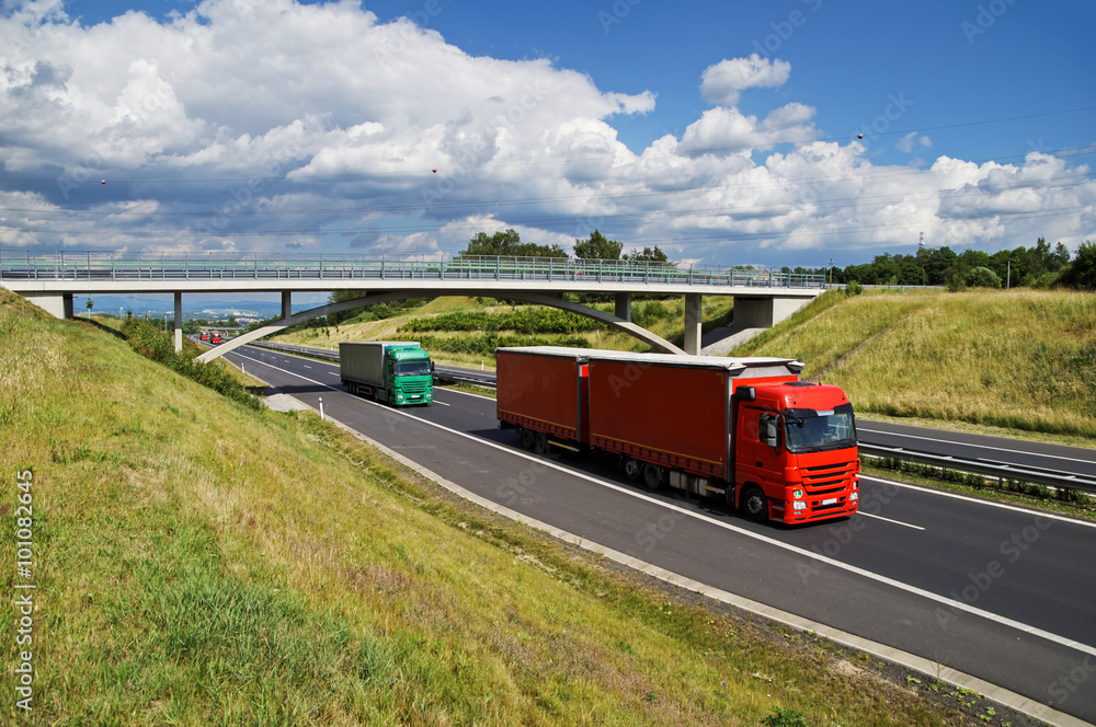 Trucks on asphalt highway goes under the concrete bridge in the countryside. View from above. Sunny day with blue sky and white clouds.