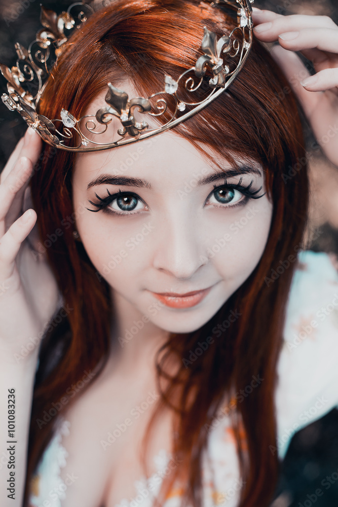 Fototapeta premium The little red-haired girl in a white dress walks in the mystical and mysterious forest.Princess with a crown standing near the tree,creative computer colors,fashionable toning