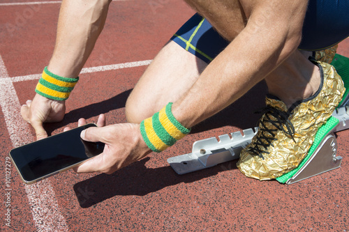 Athlete crouching at the starting line of a running track wearing Brazil colors wristbands checking his mobile phone