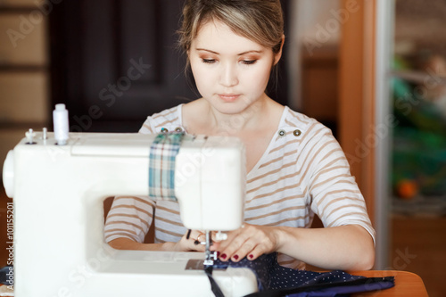 Fotografie Young woman sewing with sew machine at home while sitting by her working place