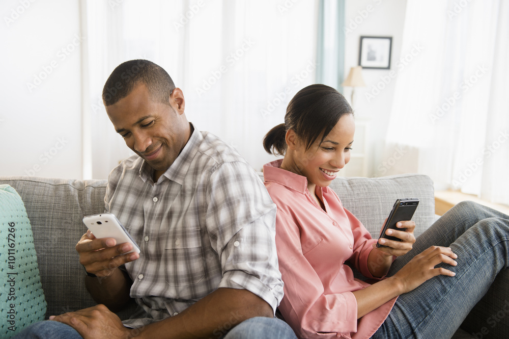 Couple using cell phones back to back on sofa Couple using cell phones back to back on sofa