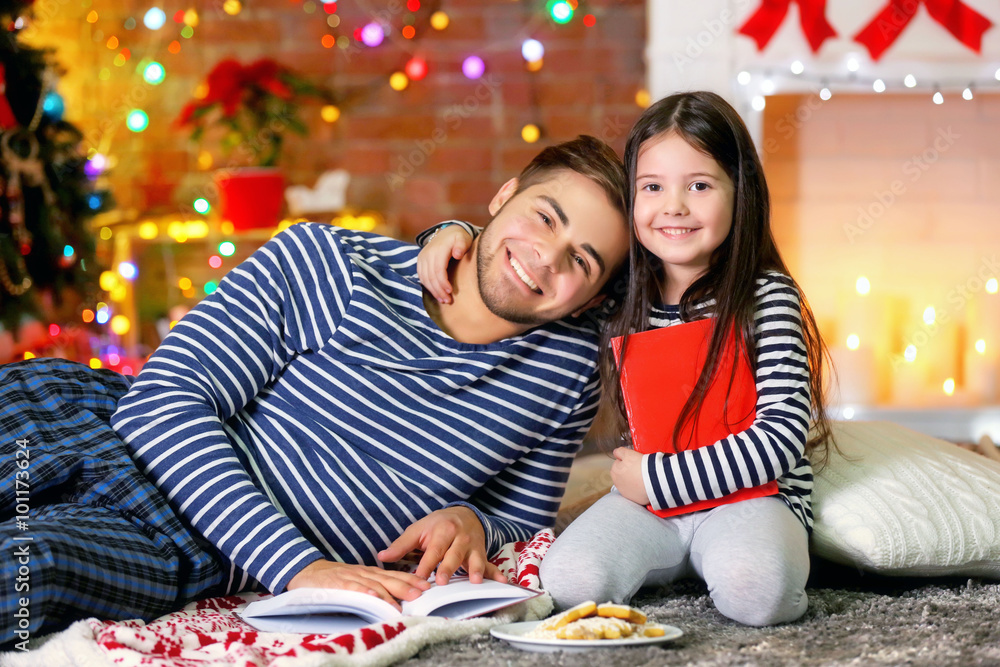 Fototapeta premium Older brother with little sister reading books and eating cookies in Christmas living room