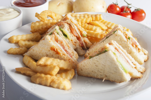 Club sandwiches with grilled chicken, tomato, lettuce, cheese, cucumber and dressing sauce, served on a white plate with french fries.