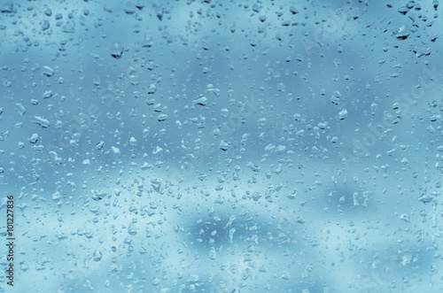 Droplets of rain on a window against a blue sky backdrop, toned blue