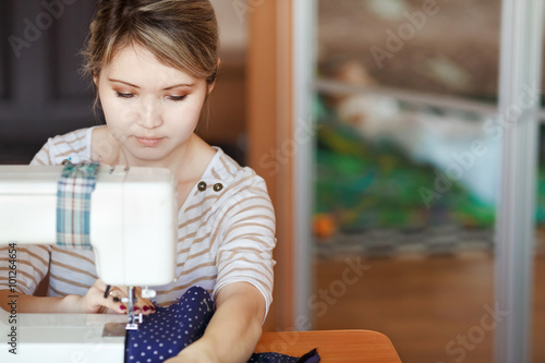 Fotografie Young woman sewing with sew machine at home while sitting by her working place