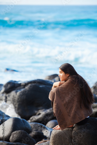 Teen girl wrapped in towel sitting on rocky beach