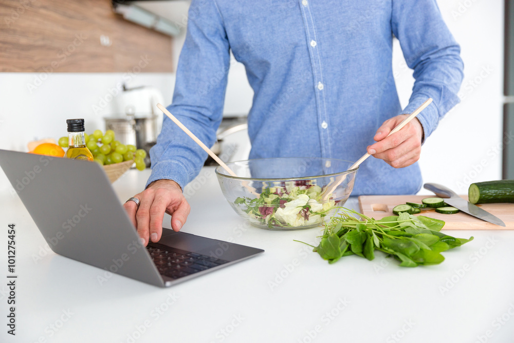 Man using laptop and making salad in glass bowl