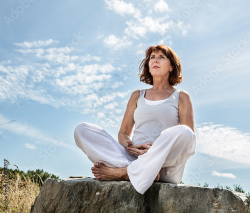 breathing outdoors - beautiful middle aged woman sitting on a stone in yoga lotus position, wearing white, seeking for balance over summer blue sky,low angle view. photo