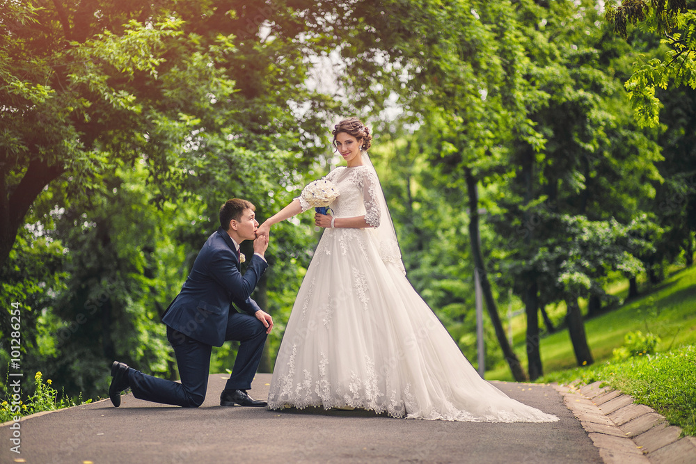 The groom down on one knee and kisses the hand of the bride in the park