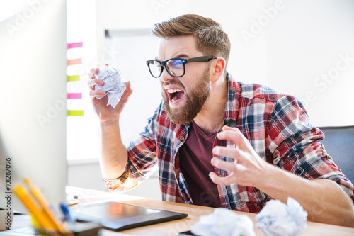 Angry crazy designer yelling and crumpling paper on his workplace photo