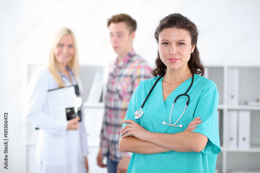 Beautiful young smiling female doctor standing at hospital with doctor and patient on the background. Medical concept. Beautiful young smiling female doctor standing at hospital with doctor and patient on the background. Medical concept.