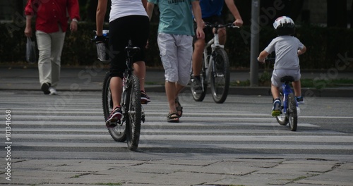 Woman And The Kid In Helmet Ride On The Bicycles Across The Crosswalk Other Bicyclers And Pedestrians Move On The Crosswalk Car Moves By Paved Auto Road Close view