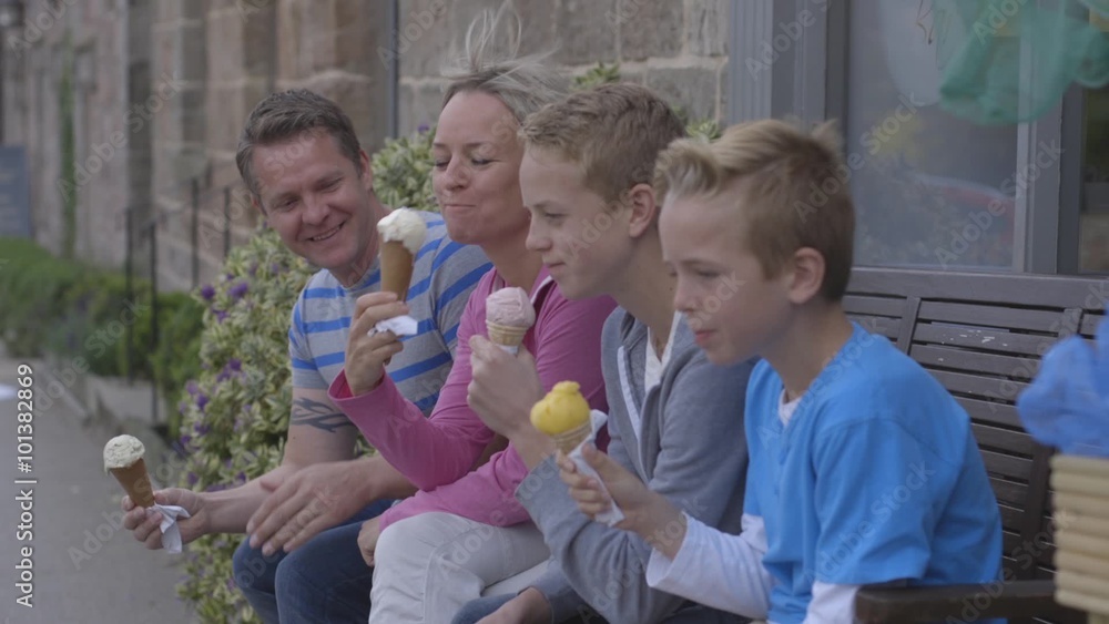 Family of four enjoying ice-cream sitting on a bench outside. They are wearing casual clothing and laughing. Family of four enjoying ice-cream sitting on a bench outside. They are wearing casual clothing and laughing.