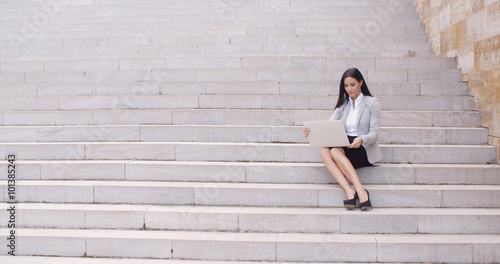 Pretty young female worker with brown hair sitting alone on marble staircase with laptop outdoors