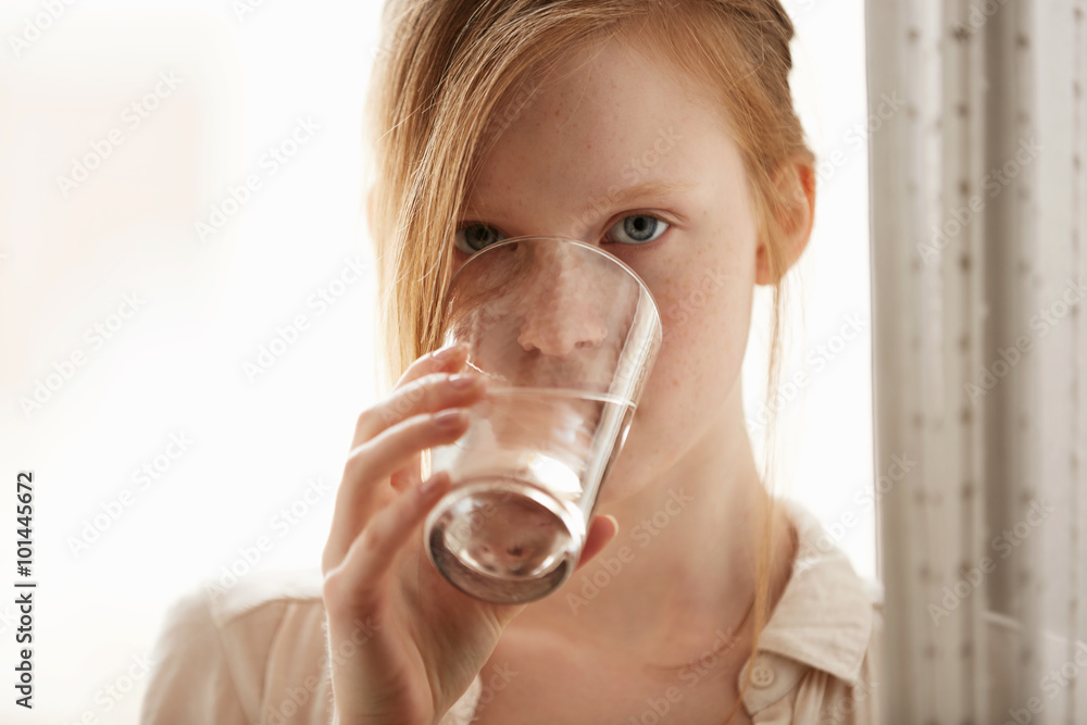 Young healthy redhead and freckled girl drinking a glass of water in bright bedroom.