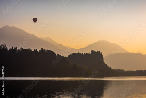 Silhouette of Bled Castle on a Lake at Sunrise with Mountains and Hot Air Balloon Flying