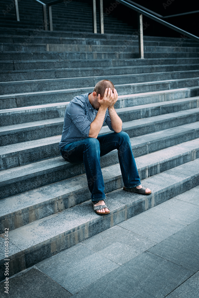 Despaired young man covering his face with hands sitting on stairs of big building. Feelings of sadness, despair and tragedy