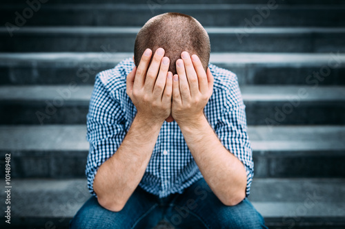 Outdoor portrait of sad young man covering his face with hands sitting on stairs. Selective focus on hands. Sadness, despair, tragedy concept photo