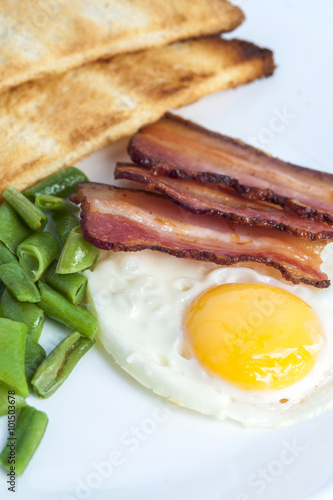 Fried egg, bacon, green beans and toasts on light background. English breakfast. Vertical view. Focus on egg and bacon.