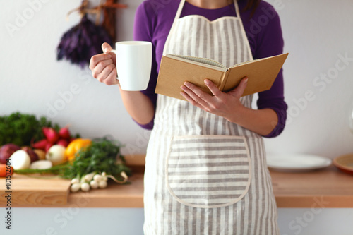 Konstfotografi Young woman reading cookbook in the kitchen, looking for recipe