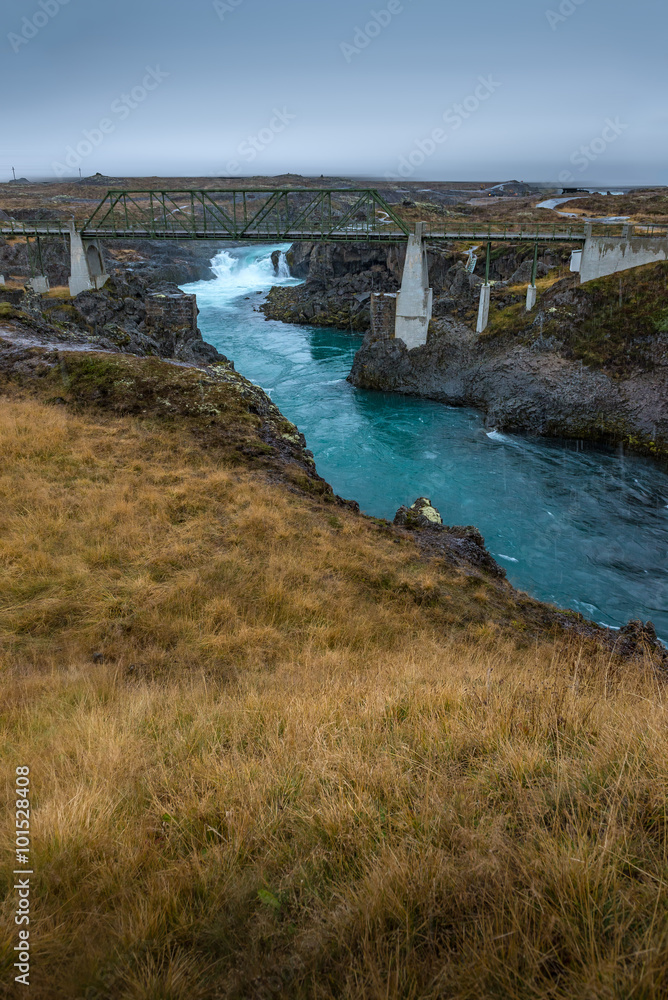 Turquoise stream and metal old bridge with yellow plant field in cloudy day Autumn season Iceland