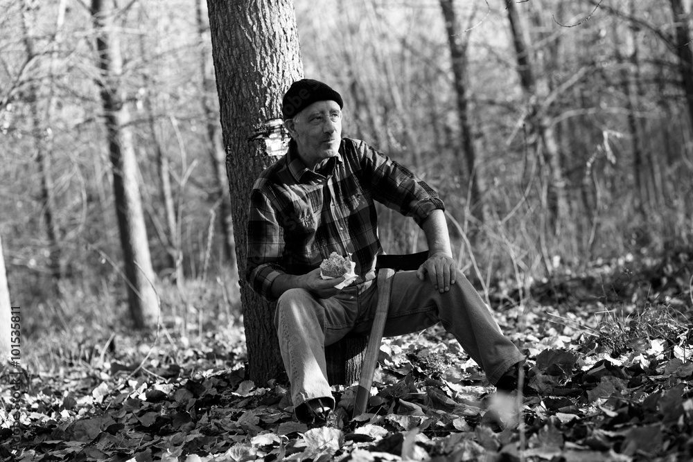 Portrait of senior lumberjack in forest sitting on a tree stump. He is resting and having lunch break. Black and white.