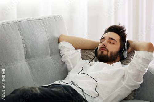 Young man listens music with headphones on grey sofa in the room