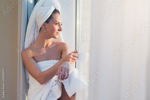 Romantic woman sitting before window and admiring sunrise or sunset with towel on her head body after bath.