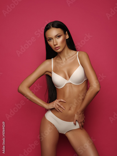 Beautiful model with dark straight hair, almond-shaped eyes and bronzed skin is posing the white basic underwear collection in the studio on the bright pink background © maksimvostrikov